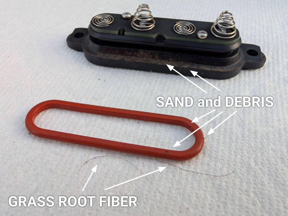 Before cleaning: A detailed close-up of a black rubber O-ring heavily coated in fine dirt and debris, with two distinct, thin strands of dried lawn roots tangled around it